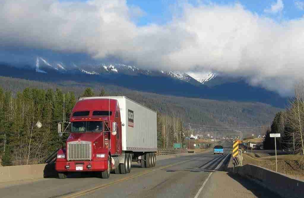 Red semi-truck driving on a highway with mountainous scenery in the background.
